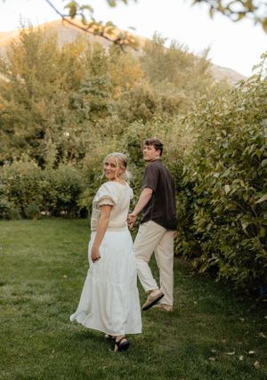 A couple walks hand in hand through a lush green garden, glancing back with a warm smile.