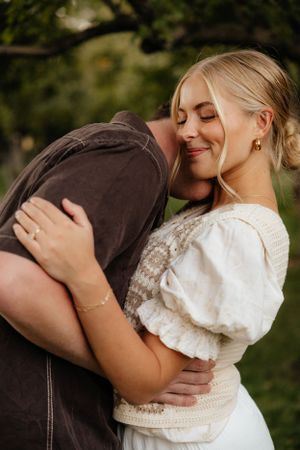 Blonde woman smiling with eyes closed while being embraced by partner outdoors in golden light.