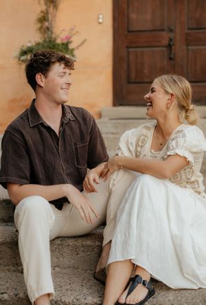 Couple sitting on stone steps laughing together, man in brown shirt and woman in white boho dress.