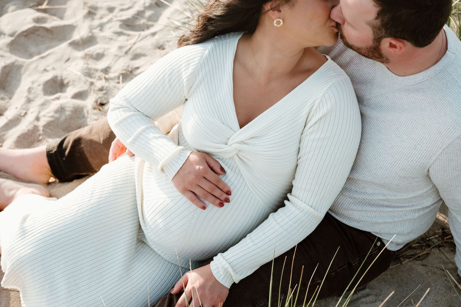 Intimate maternity photo of couple in white sweaters sitting on beach.