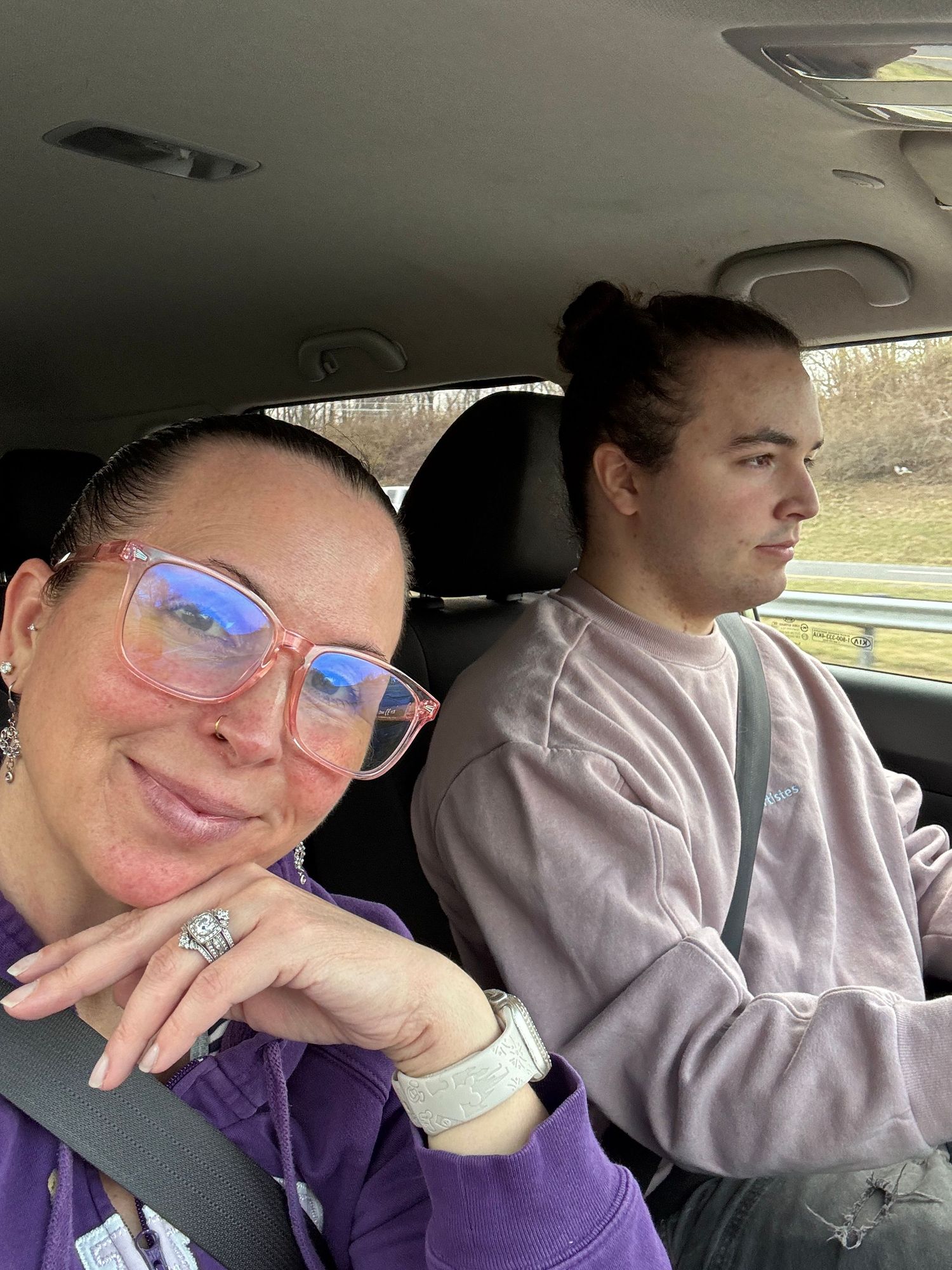 Two people sitting in a car interior, with one wearing a purple sweater and clear glasses while the other sits in passenger seat.