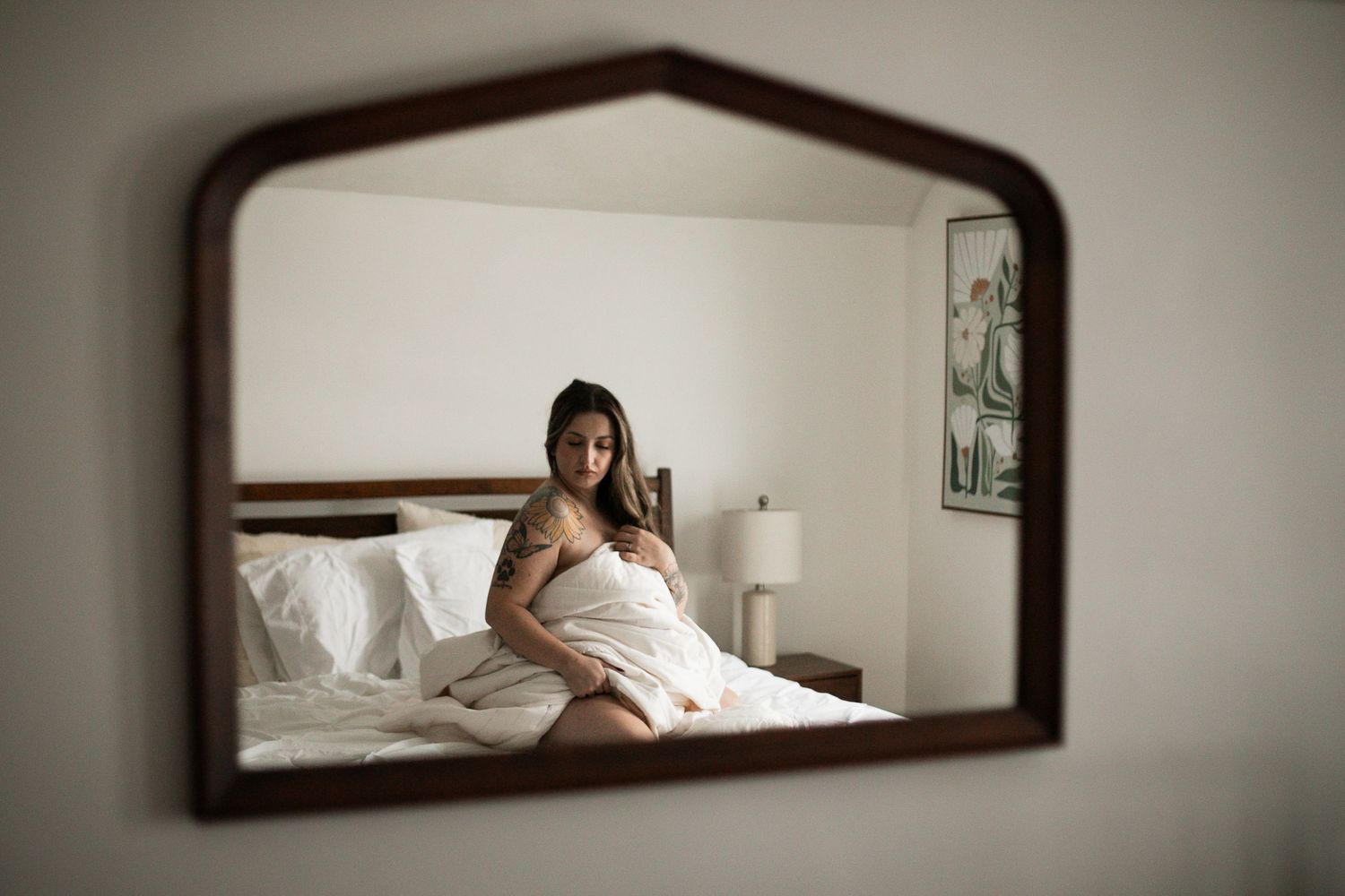 A person in red lingerie with visible tattoos poses thoughtfully on a white bed near artwork on the wall.