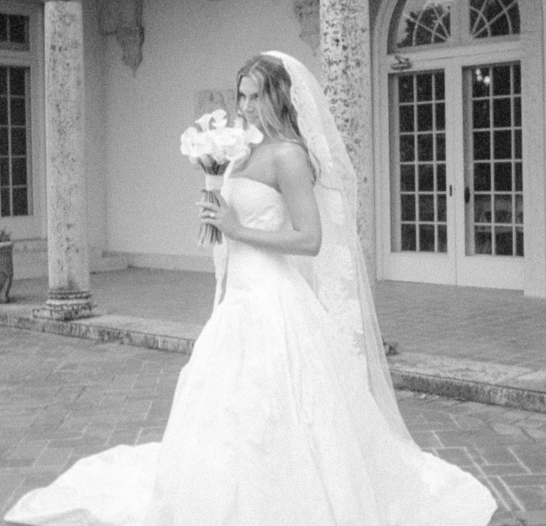 A bride in a flowing white wedding gown and long veil holds a bouquet while standing on a stone patio near arched windows.