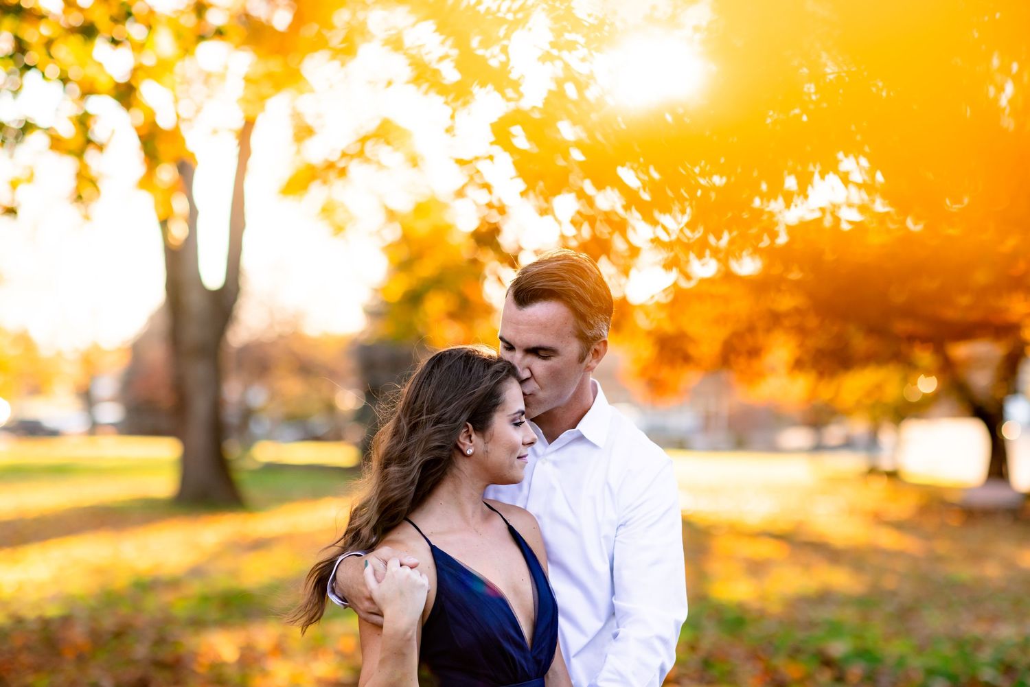 Couple embraces in romantic autumn setting with golden sunlight filtering through yellow fall foliage.