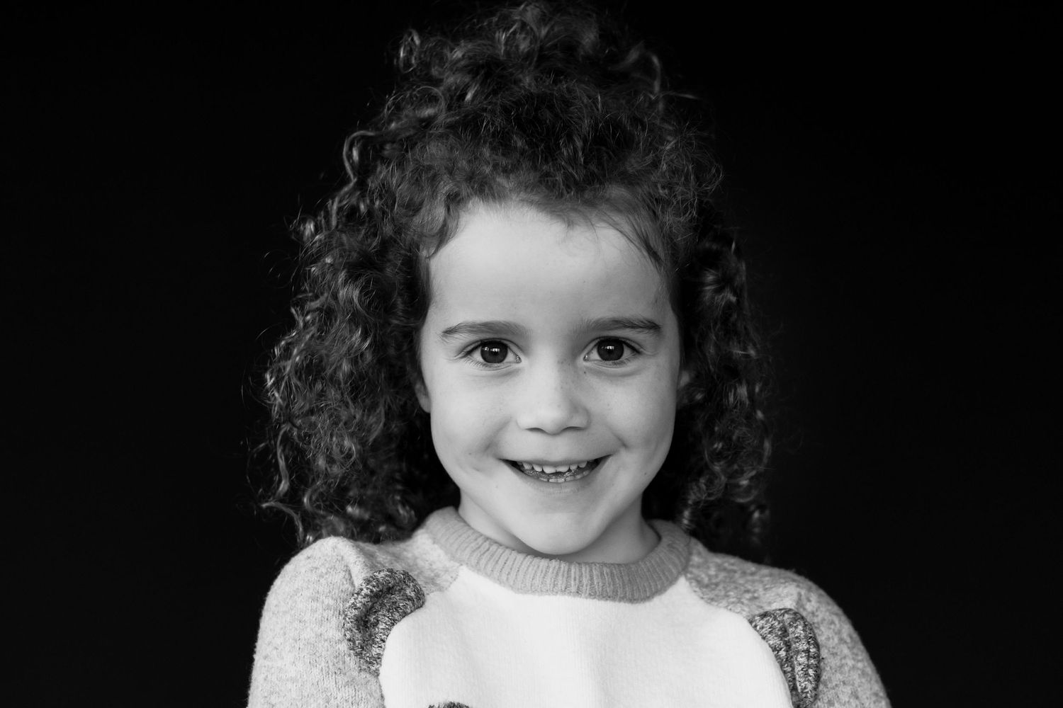 Young girl with curly dark hair smiling sweetly in a black and white portrait photograph.