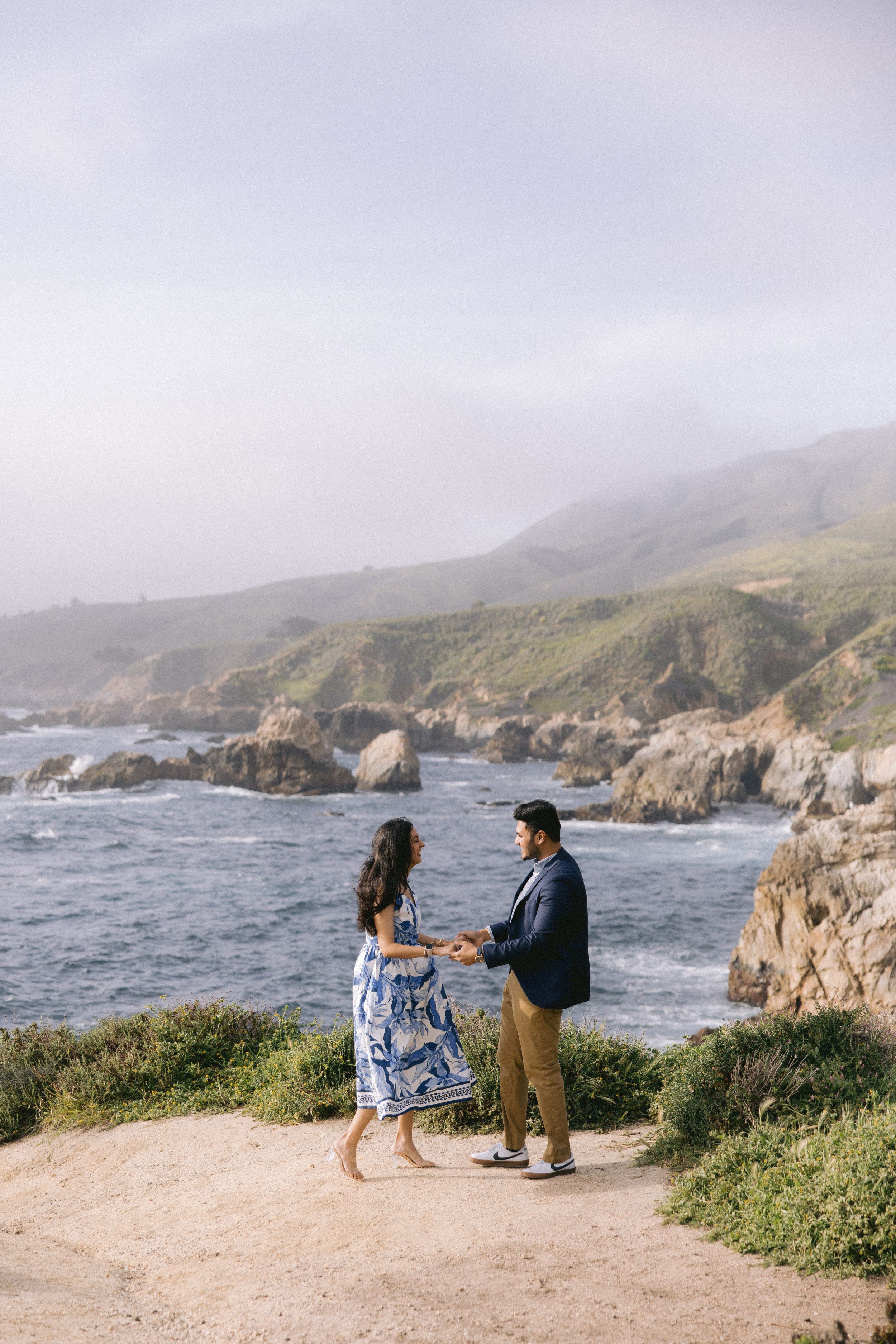 A couple stands on a coastal cliff overlooking the rocky Pacific shoreline on a misty afternoon.