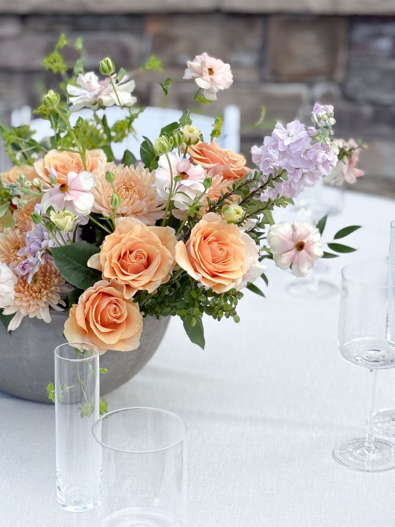 Elegant floral centerpiece with peach roses, white flowers, and greenery arranged on a white tablecloth with glasses.