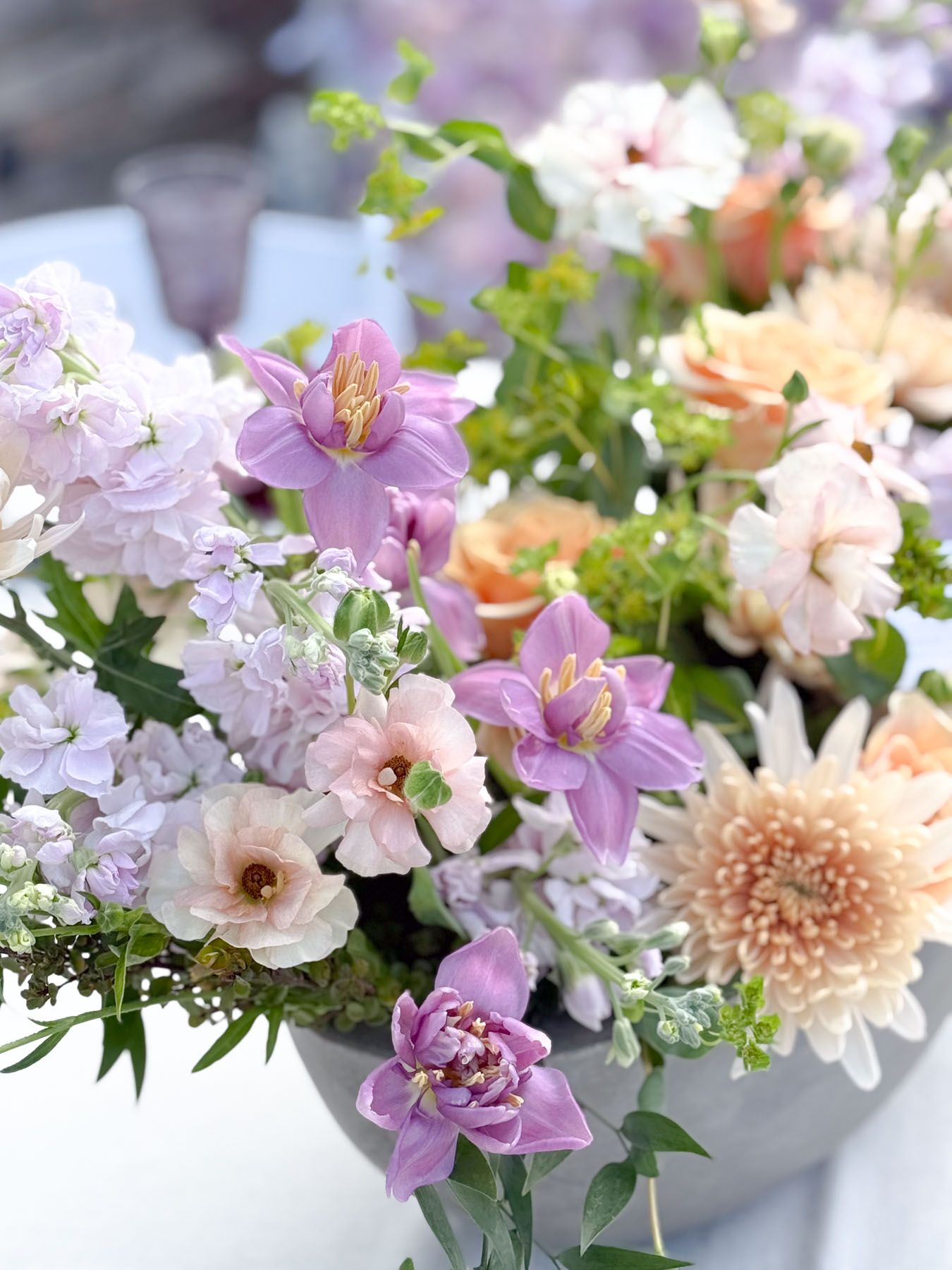 Close-up of a delicate floral arrangement featuring purple lilies, pink carnations, and peach-colored blooms in soft focus.