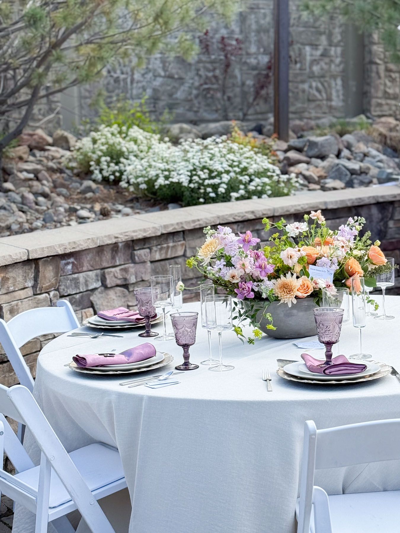 Elegant outdoor table setting with white linens, purple plates, and a colorful floral centerpiece against a garden backdrop.