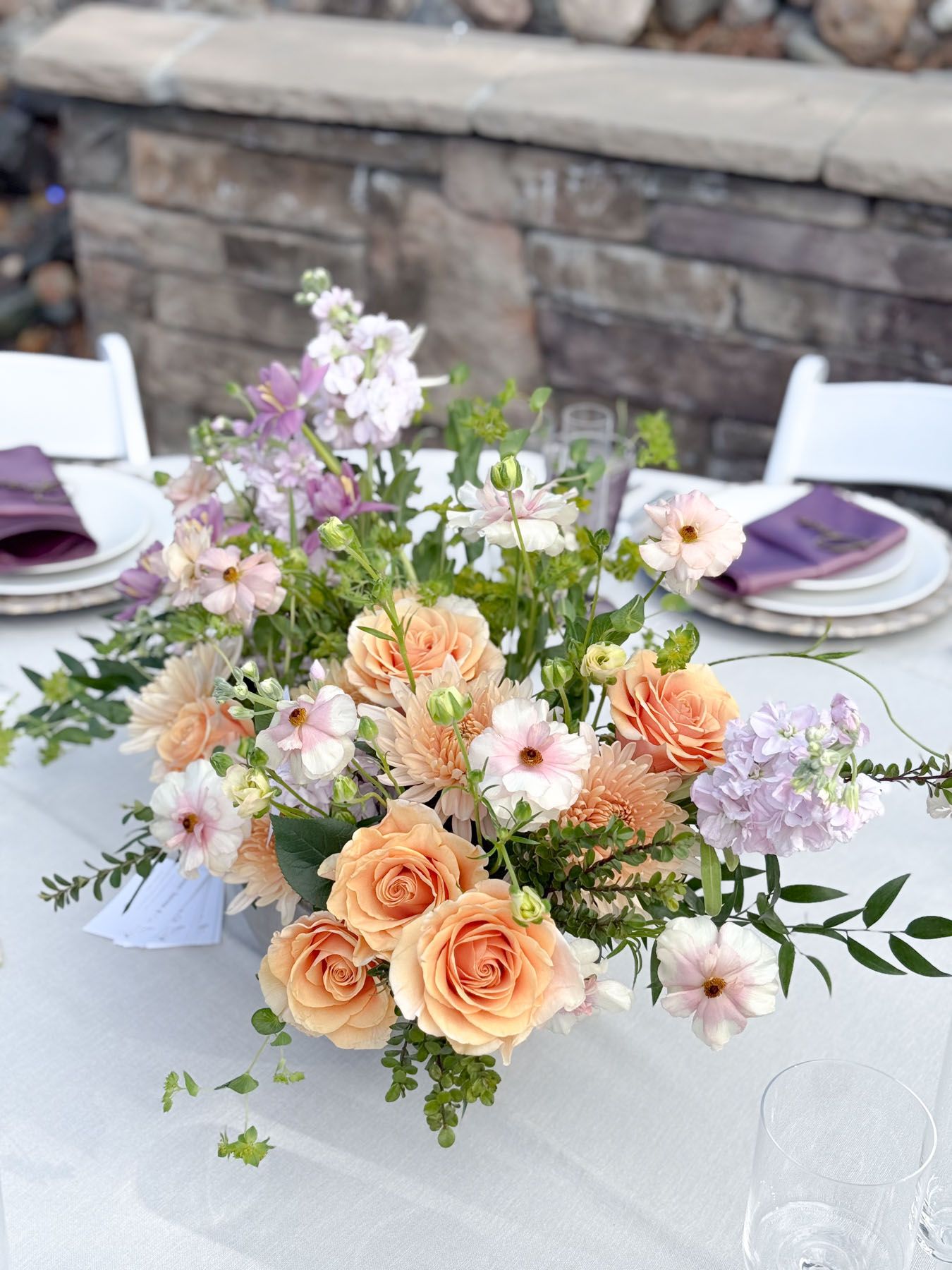A beautiful floral centerpiece with peach roses, pink flowers and greenery on a white tablecloth with purple place settings.