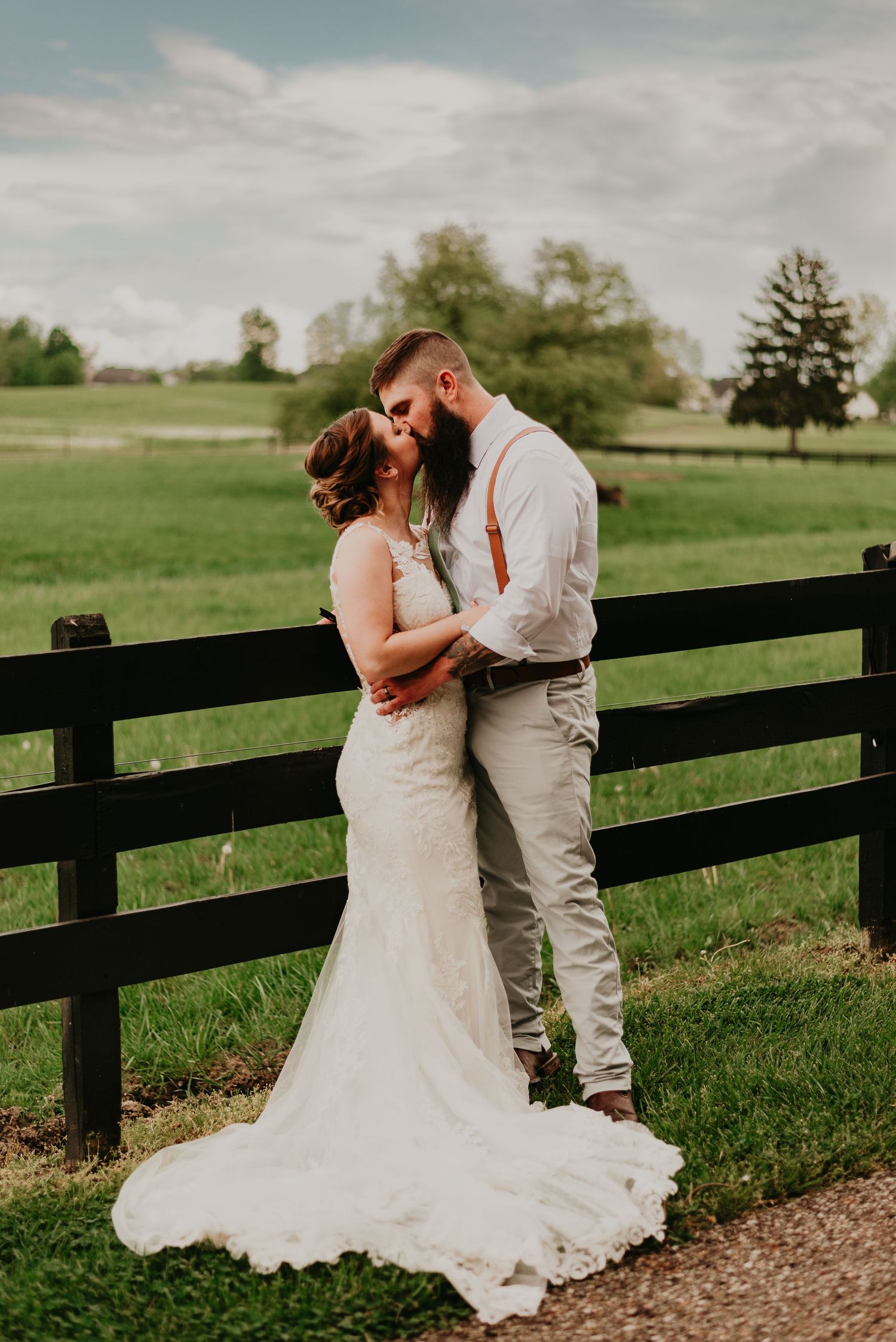 A romantic wedding couple shares intimate moments along a black fence in a lush green countryside setting.