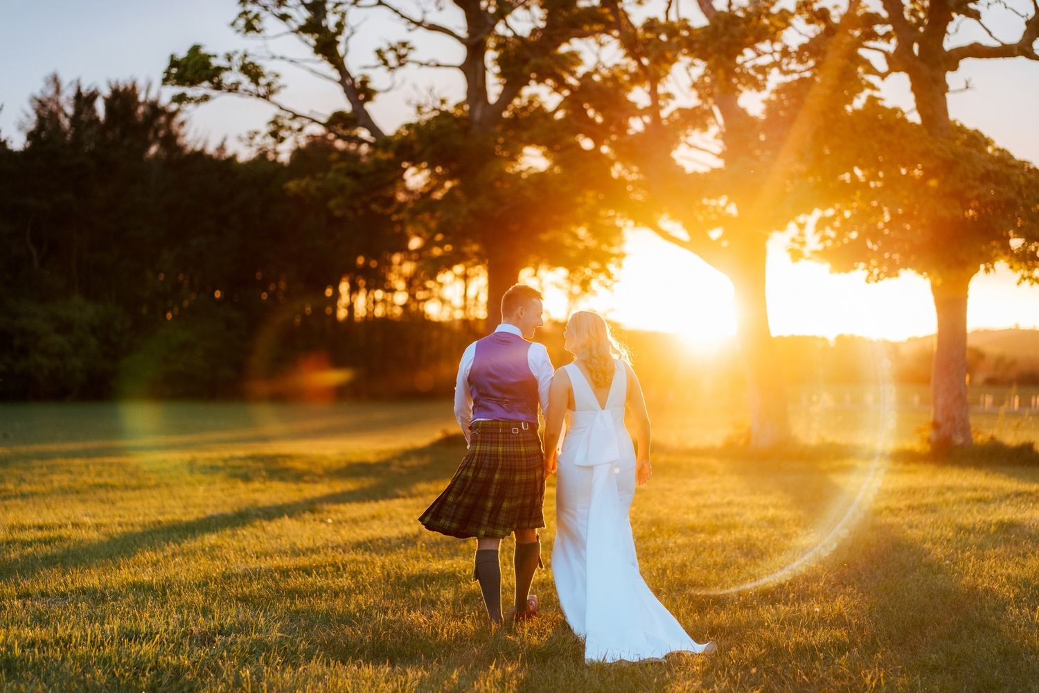 A couple in formal attire walking on a beach at sunset, casting long shadows on the golden sand.