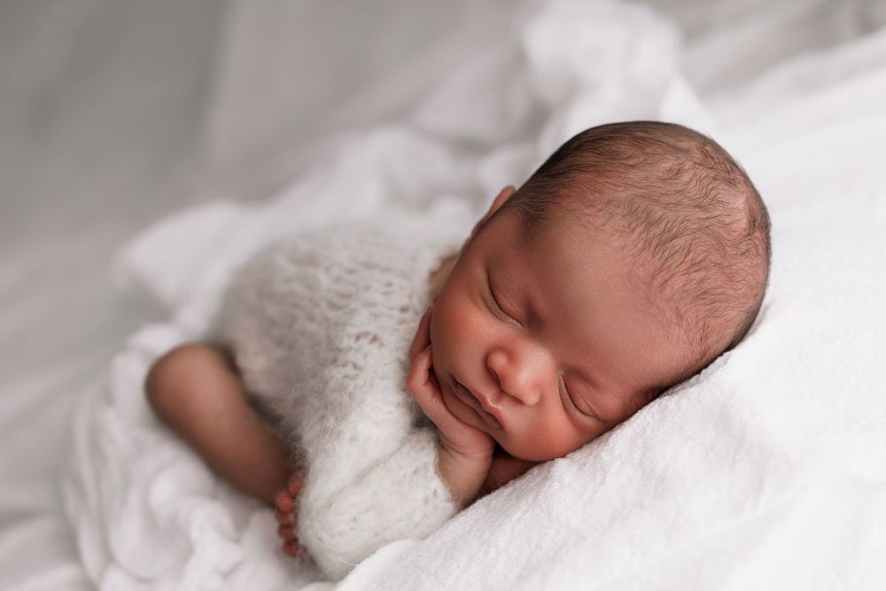 A sleeping newborn wrapped in a soft white blanket rests peacefully on white bedding during a professional photography session.