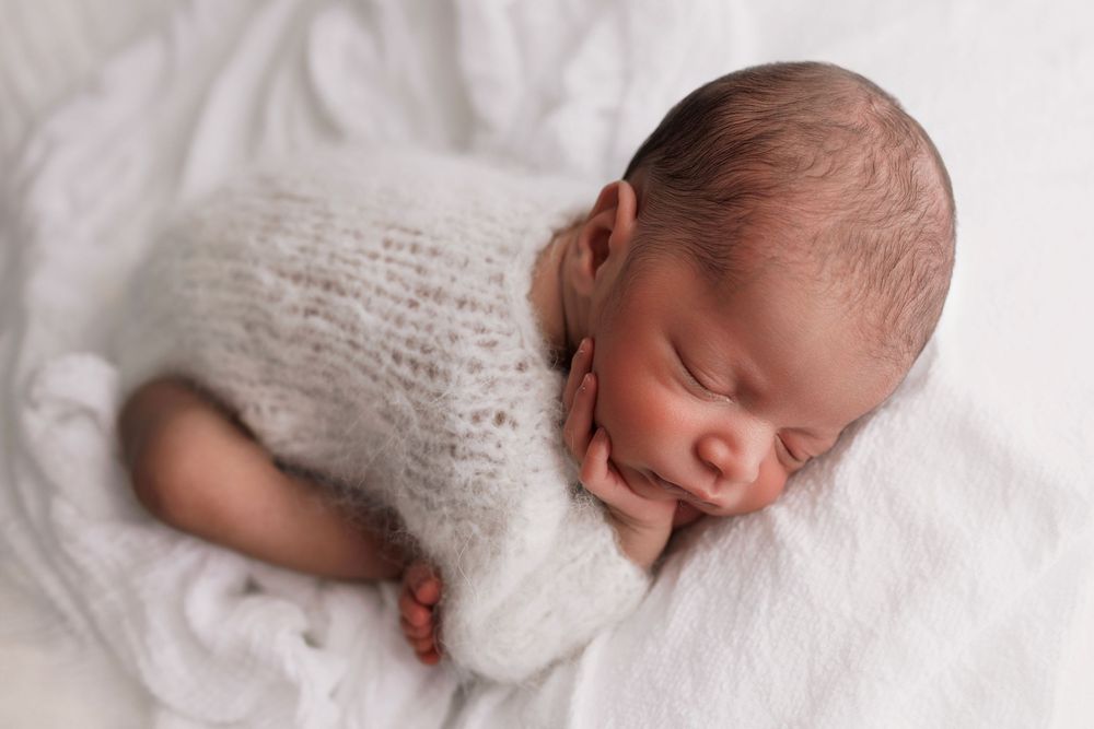 Peaceful sleeping newborn baby wearing soft white knit wrap rests on white blanket during professional photo session.