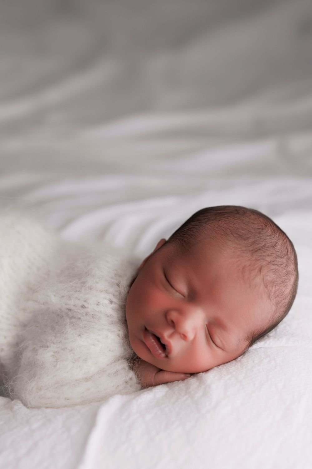 A peaceful sleeping newborn baby wrapped in white swaddle blankets against soft white bedding.