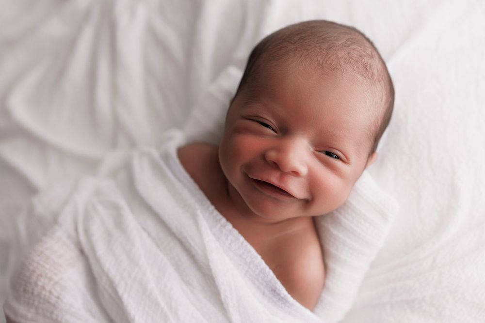 Newborn baby wrapped in white blanket smiling joyfully during professional photo session.