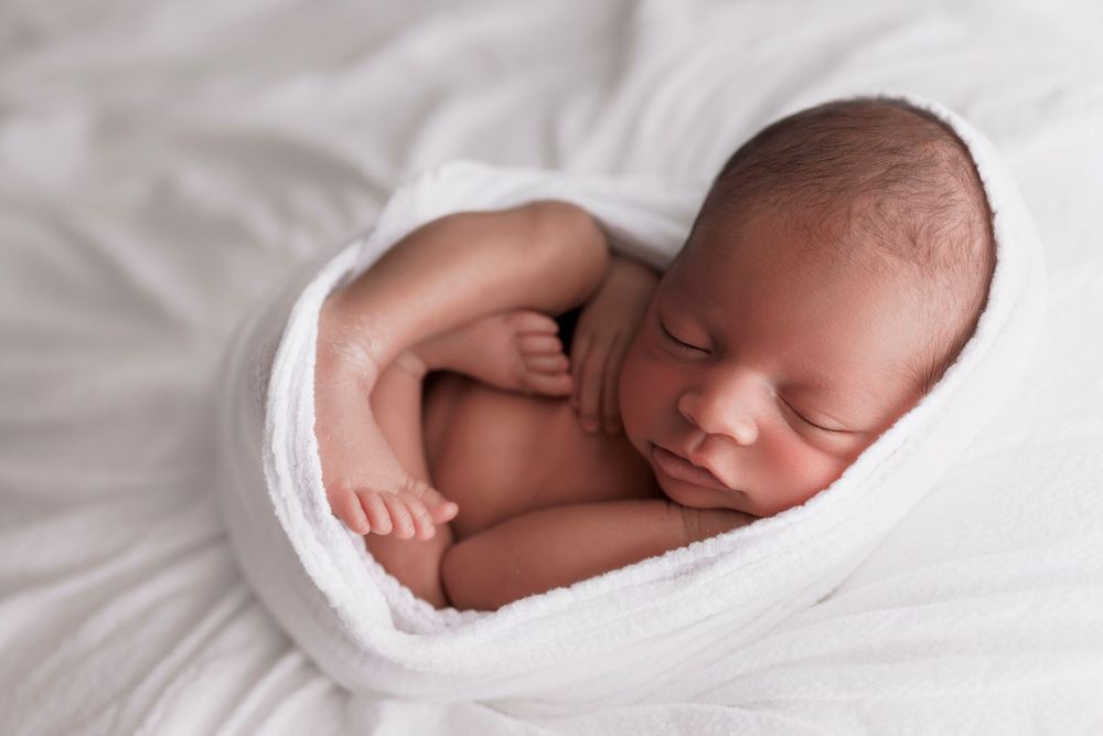 A sleeping newborn baby swaddled in soft white fabric lies peacefully against white bedding.