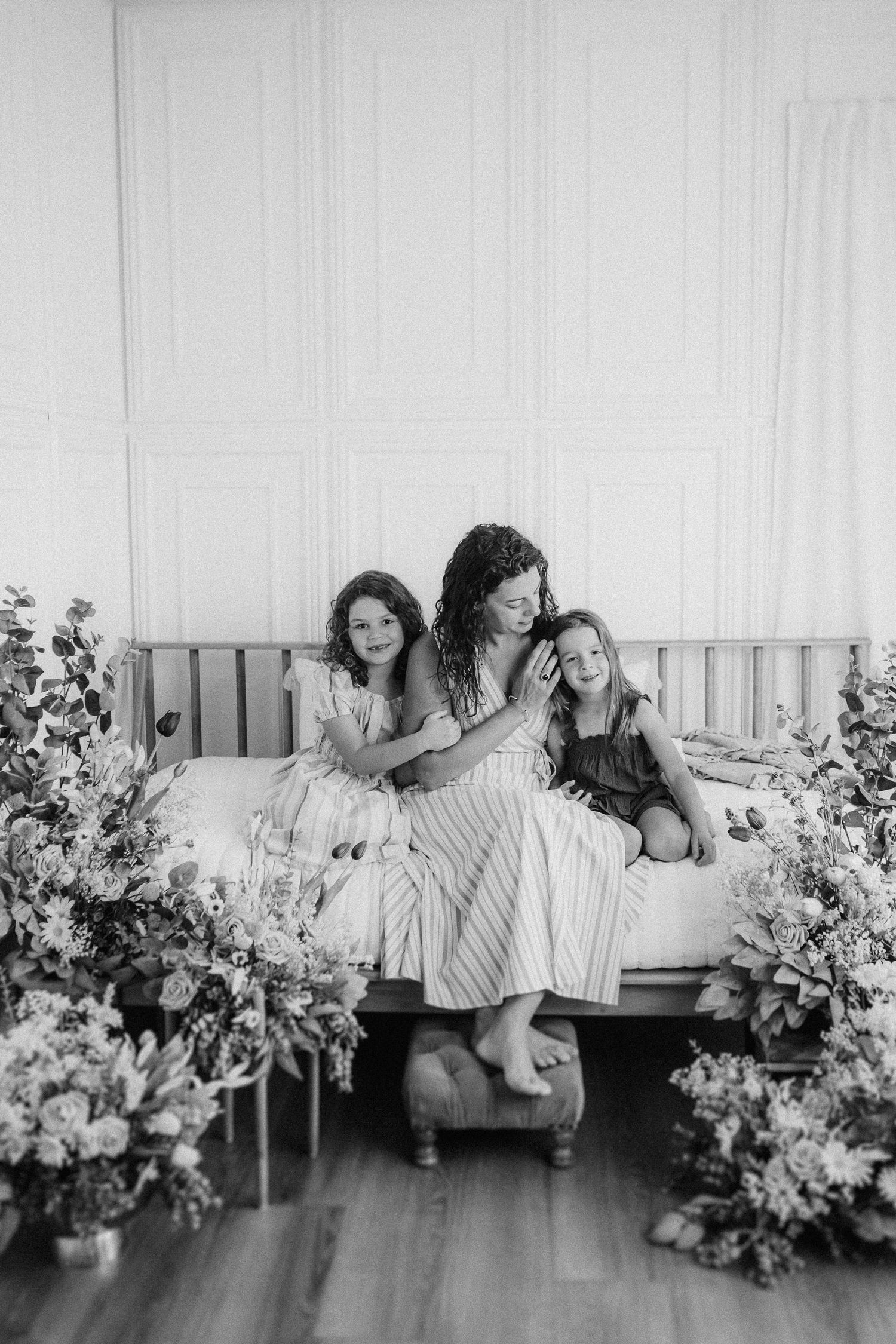 Three figures share joyful moments on a white bench surrounded by lush floral arrangements in a black and white photo.