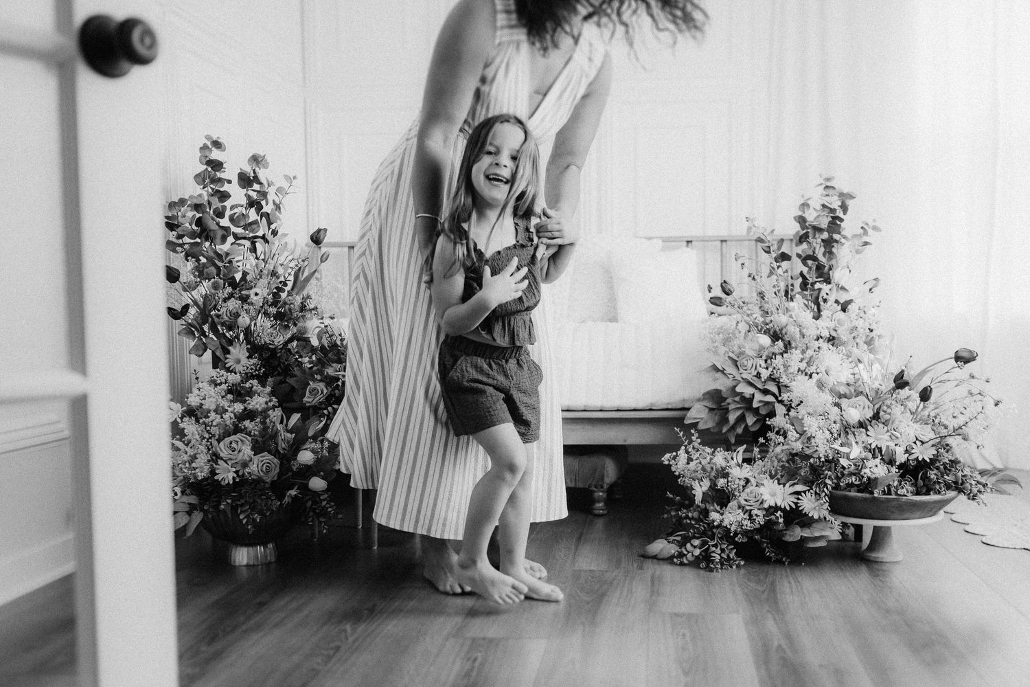 A joyful child in overalls dances and spins in front of a white wall with potted plants on a porch.