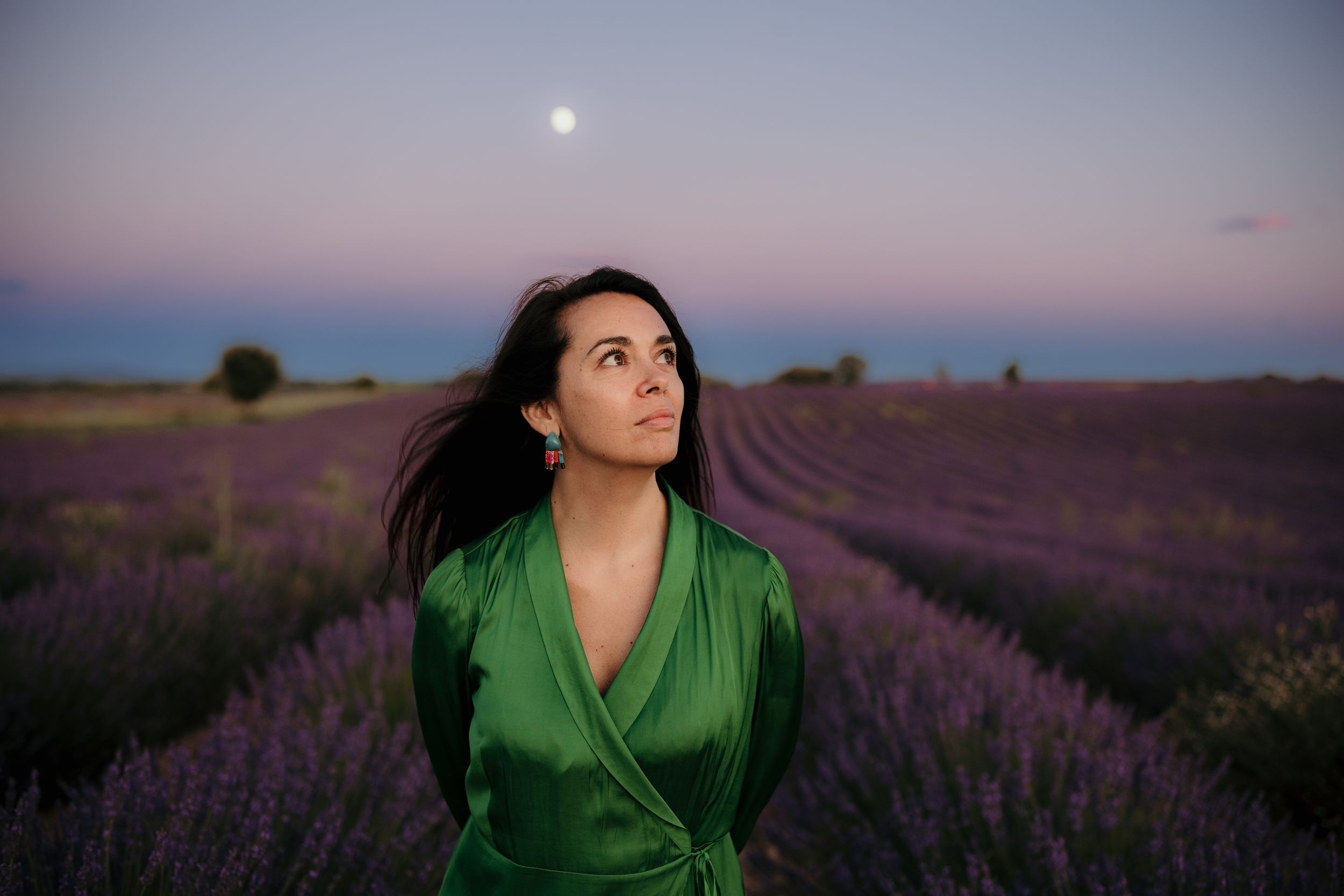 A person in an emerald green dress gazes up at the moon over a blooming lavender field at dusk.