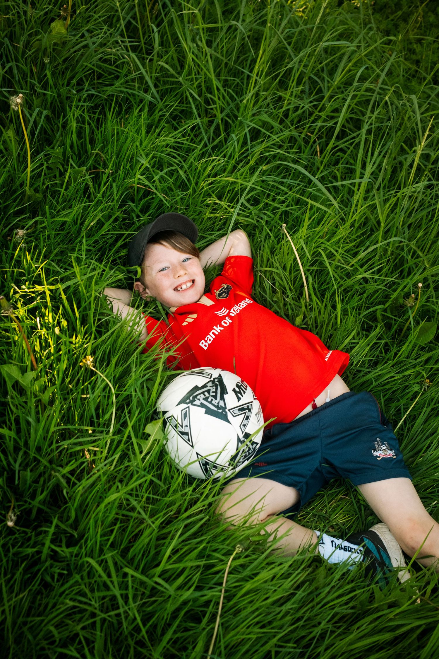 A child in a red shirt lies in tall grass with a soccer ball beside them.