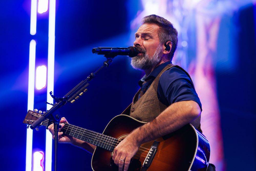 Christian artist Mac Powell of Third Day performing on stage with acoustic guitar at a concert with blue stage lighting.