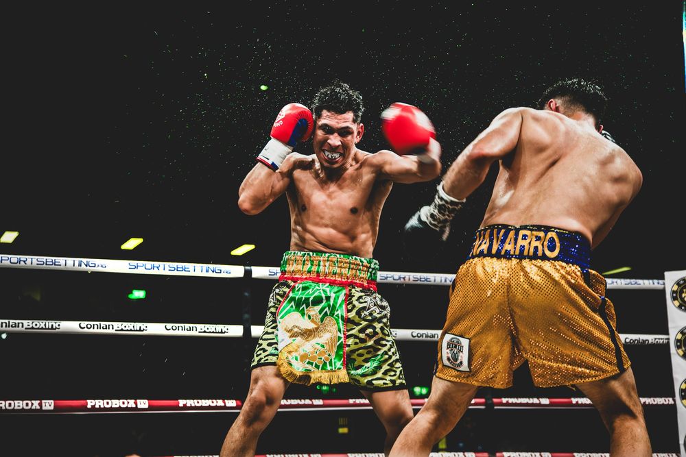 Two boxers square off in a boxing ring, one wearing colorful green trunks and one in gold trunks during an intense match.