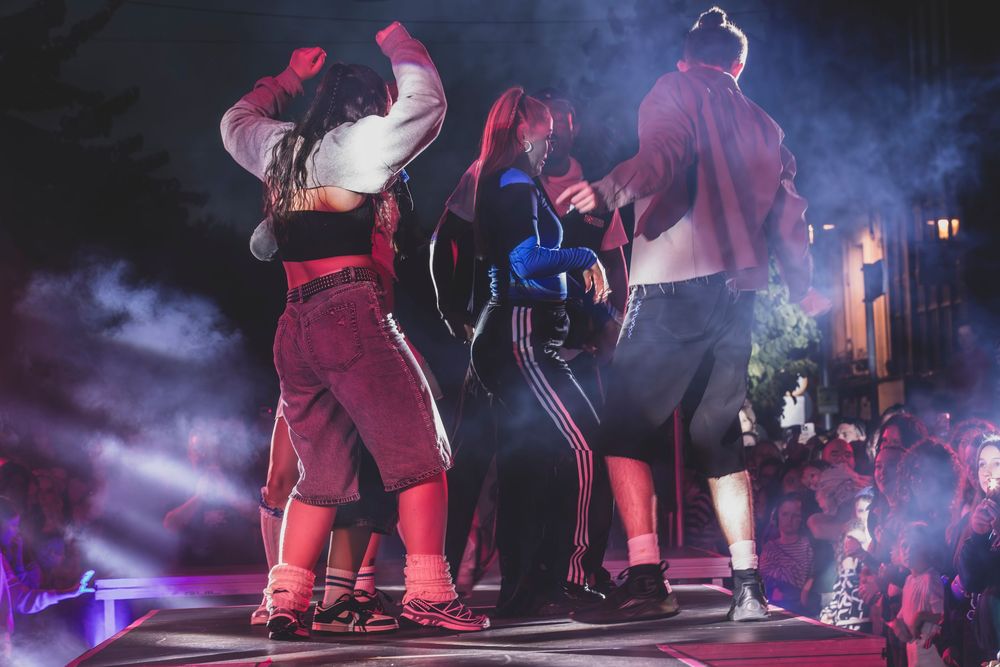 Group of dancers performing on a lit stage with colorful purple and red lighting at a nighttime concert event.