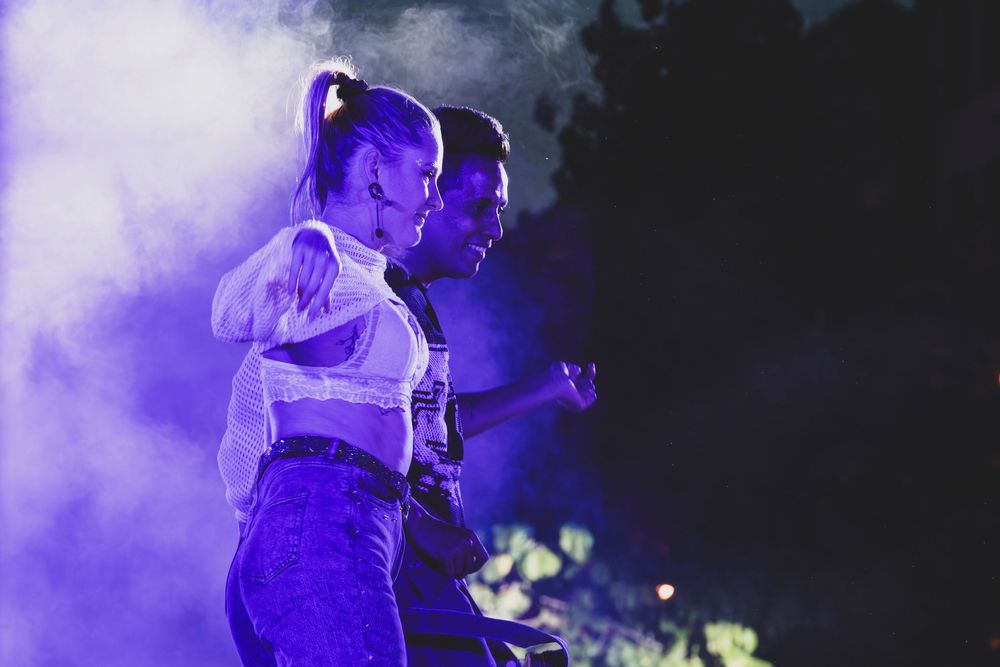 Two performers dance together on stage under purple lighting with dramatic smoke effects at an outdoor concert.
