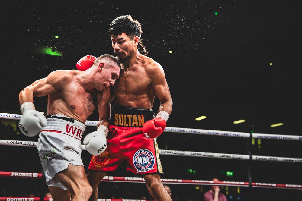 Two boxers exchange punches in a dramatic ring battle with red gloves under bright lights.