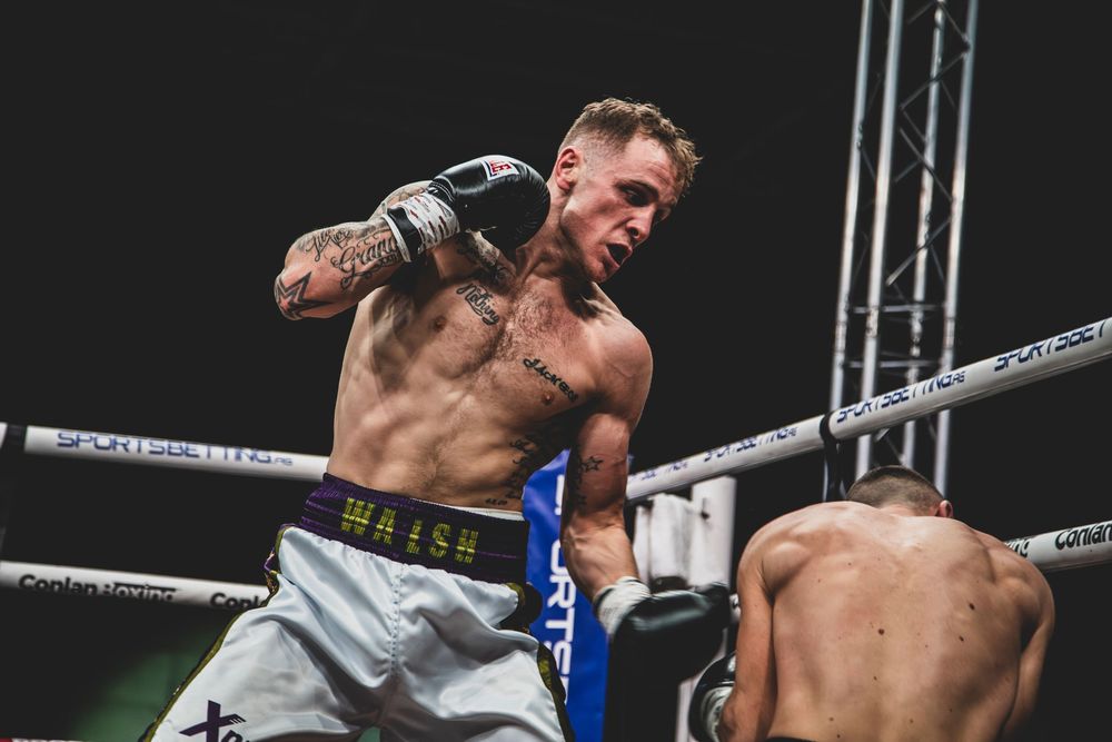 Two boxers engaged in an intense boxing match inside a professional ring with spotlights overhead.