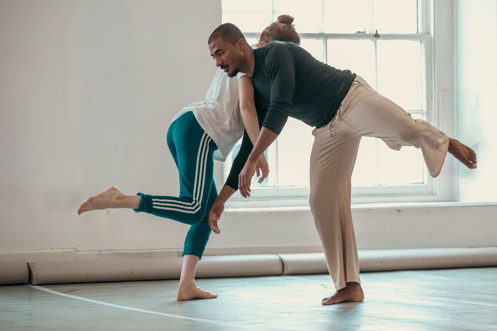 Two dancers execute a graceful contemporary dance lift in a sunlit studio with large windows.