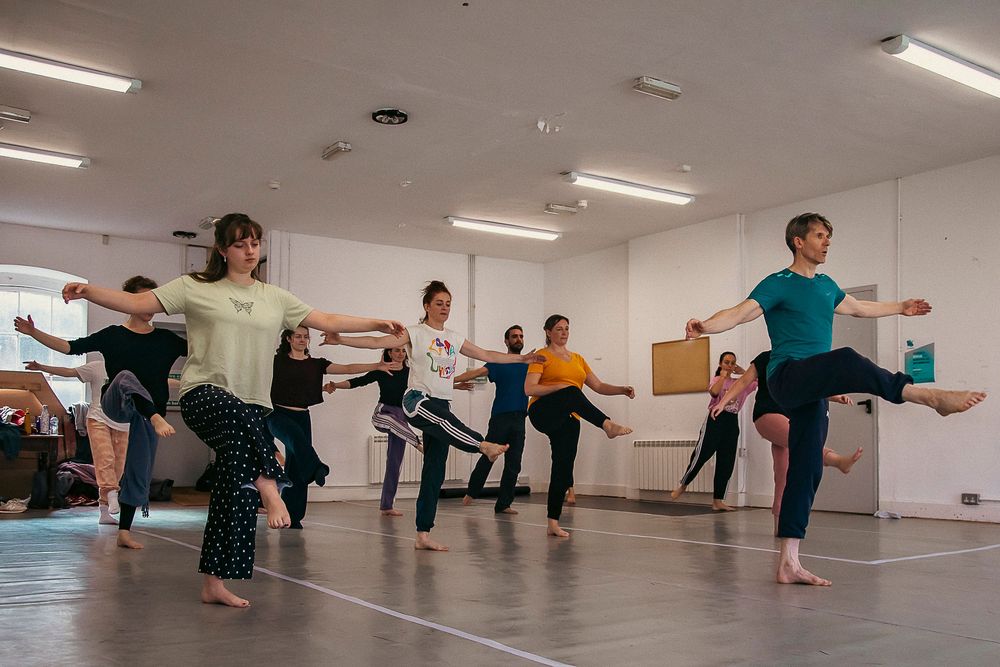 Dance students practice coordinated movements during a group class in a bright studio space with fluorescent lighting.