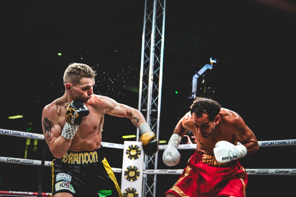 Two boxers exchange punches during an intense professional boxing match in a boxing ring under bright lights.