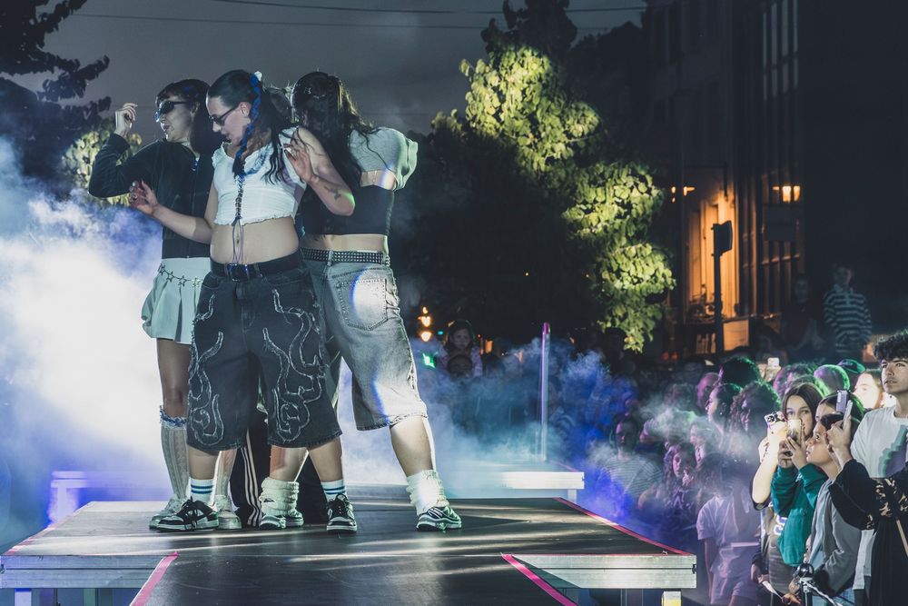 Dancers perform on a fog-filled outdoor stage at night with an audience watching in the background.