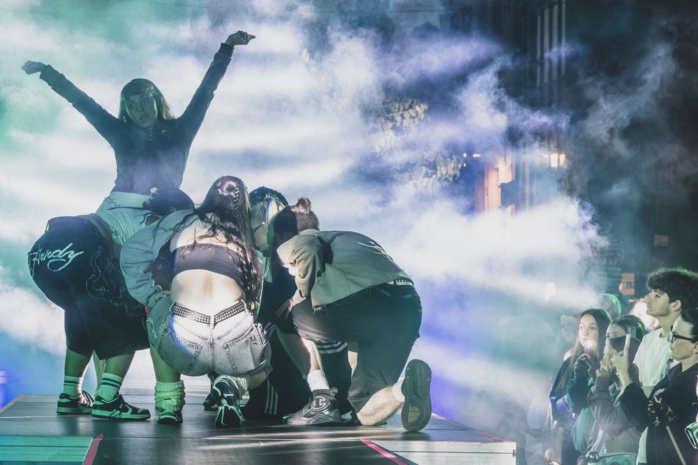 Dancers perform an energetic group routine on a smoky stage with dramatic lighting at a live concert event.