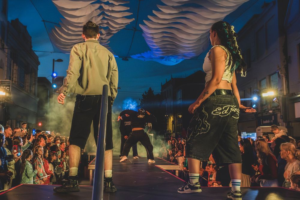 Performers on an outdoor stage at night with a crowd watching under a decorative canopy at a street festival.