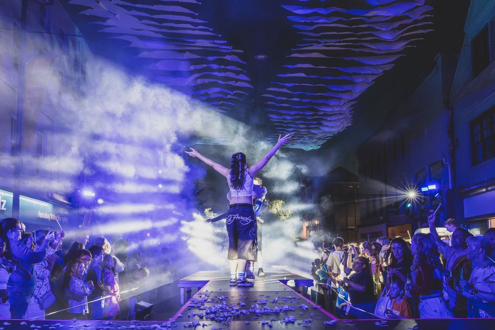 Performer on stage with dramatic blue light beams and fog at an outdoor music festival at night.