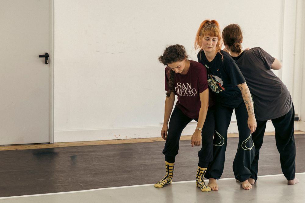 Three dancers in casual attire practice together in a dance studio with a white wall and dark floor.