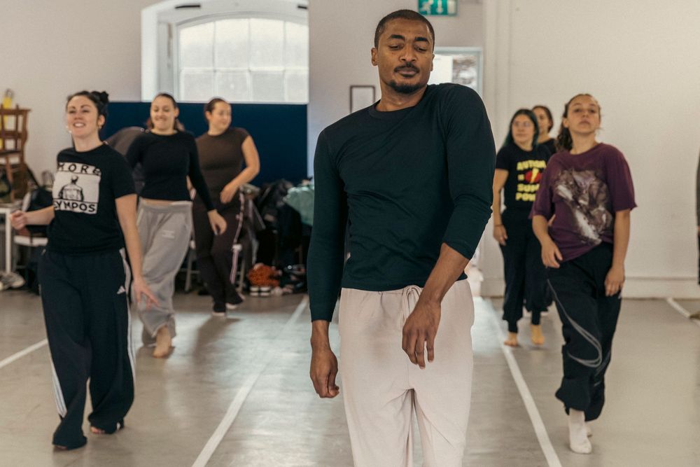Dancers in casual clothing rehearse in formation at a bright dance studio space.