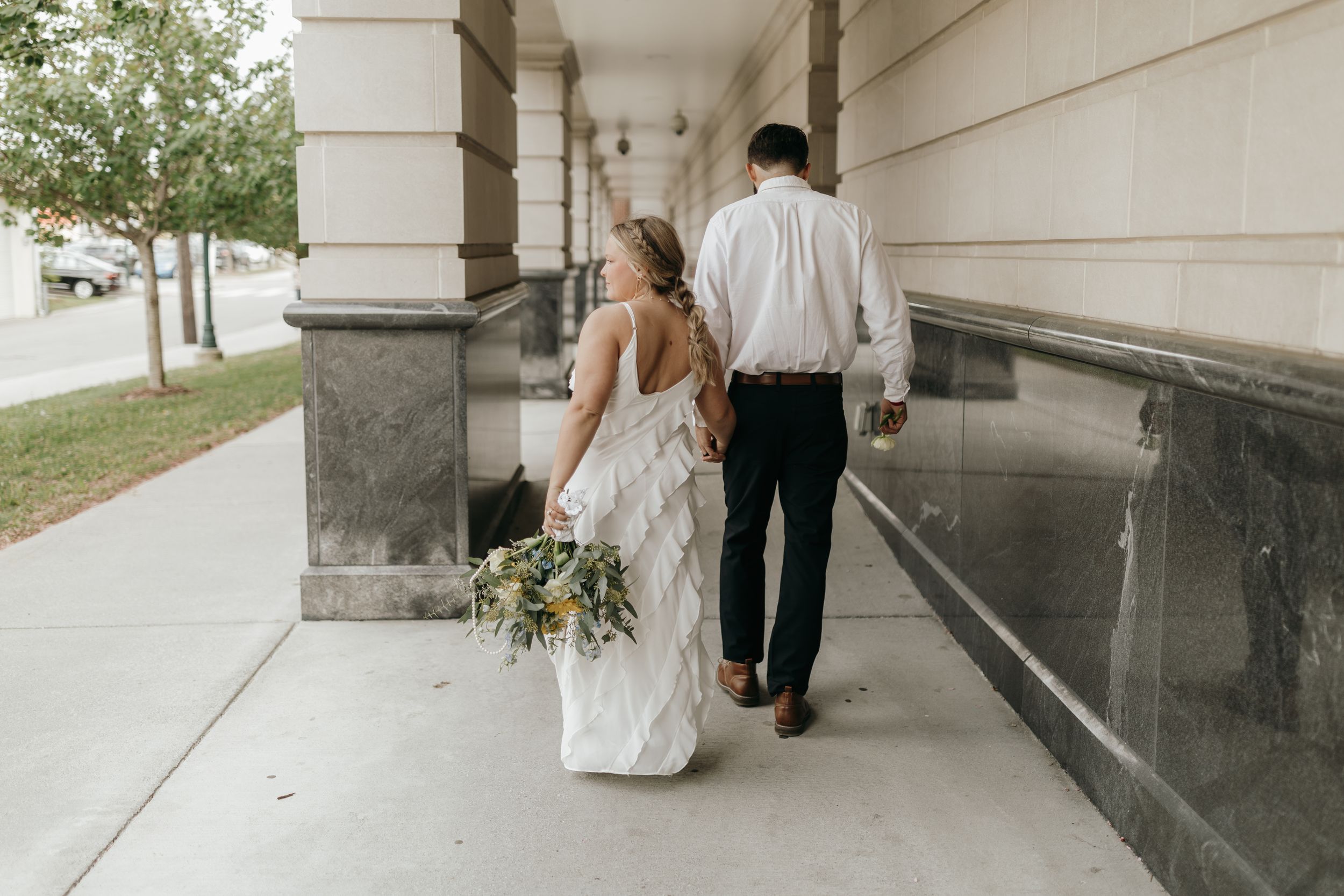 A couple walks along a pillared walkway on their wedding day, with the bride holding a bouquet of flowers.