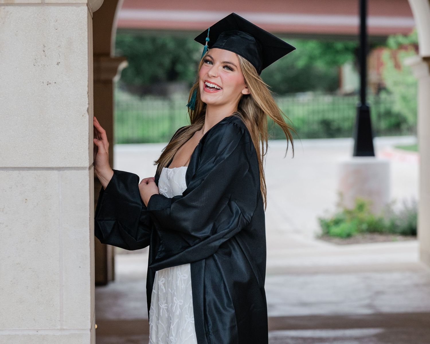 Graduate in a black cap and gown leans against a white column while smiling during a graduation photoshoot.
