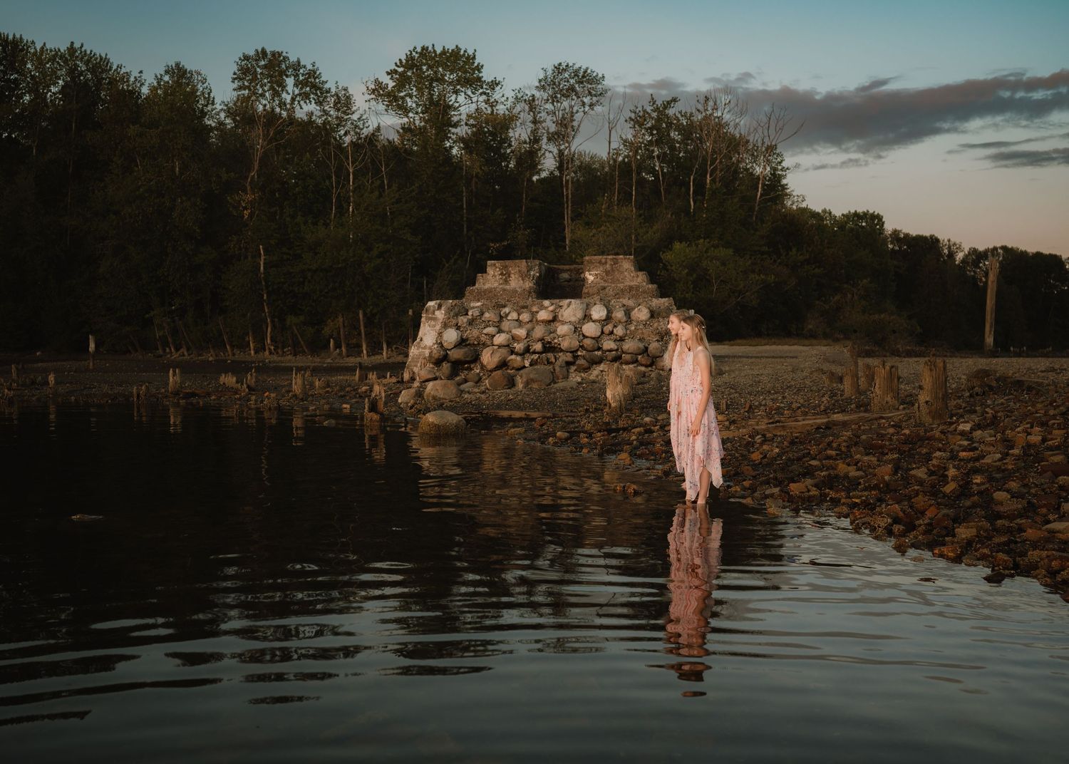 A figure in a pink dress stands in tranquil water at dusk beside a stone pier and wooded shoreline.