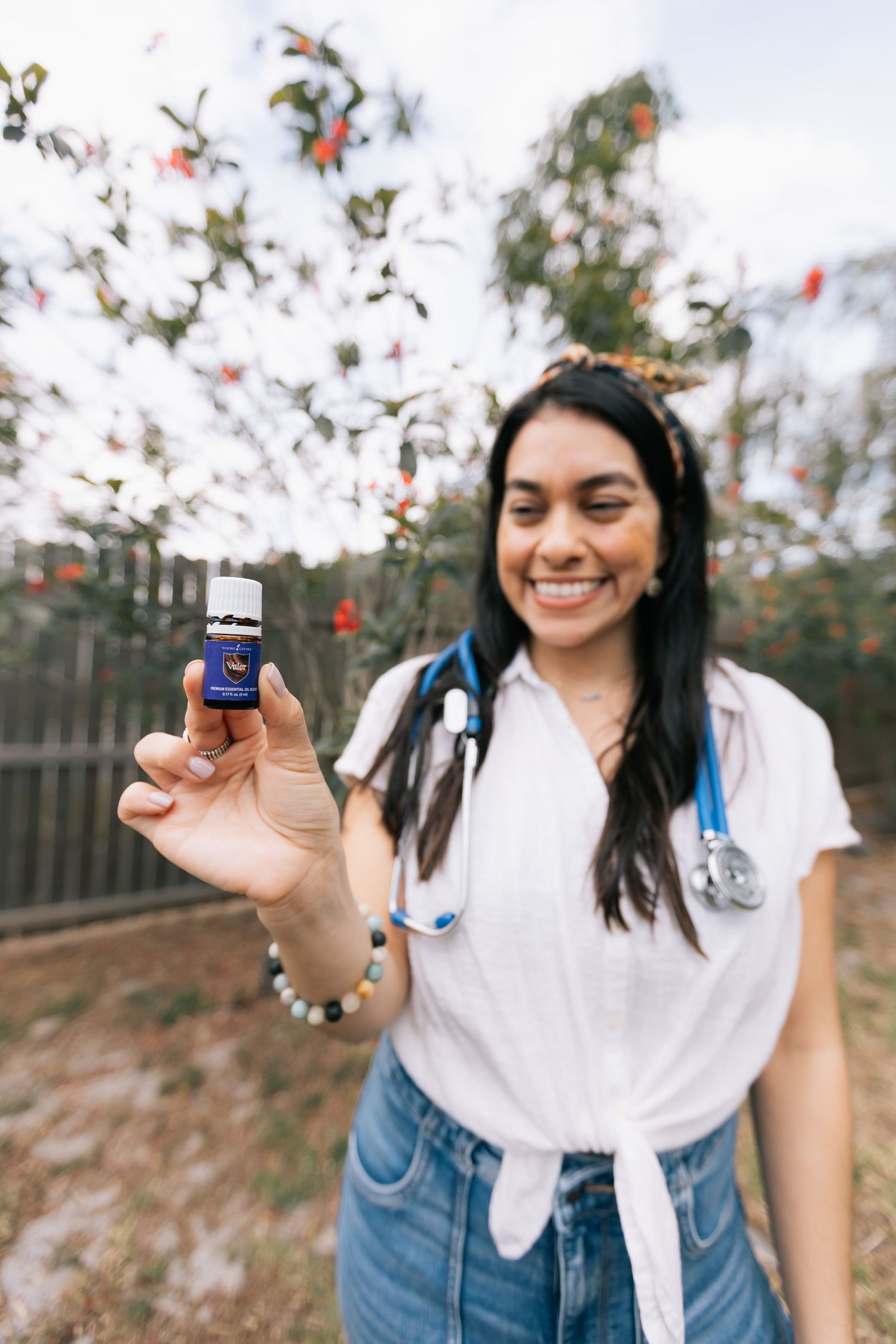 Smiling woman with a stethoscope around her neck holds up a small essential oil bottle outdoors near blooming trees.
