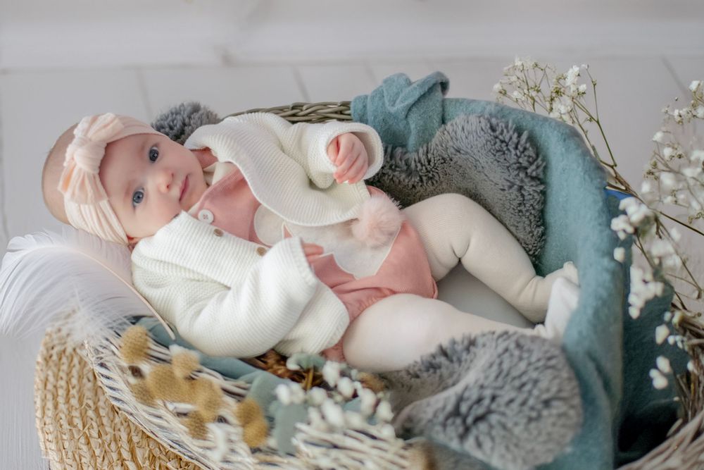 Sweet baby wearing cream sweater rests in cozy wicker basket with teal blanket and floral decor.