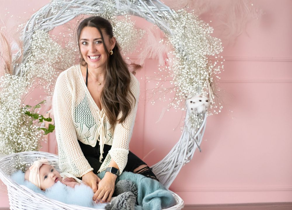 A person in a white cardigan sits beside a swaddled baby near a decorative wreath against a pink wall.