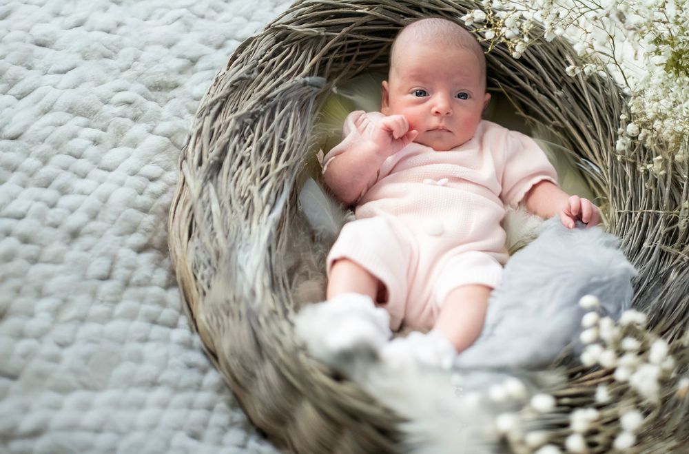 A small baby wearing pink sleepwear lies in a cozy gray woven basket with white flowers and soft textiles.