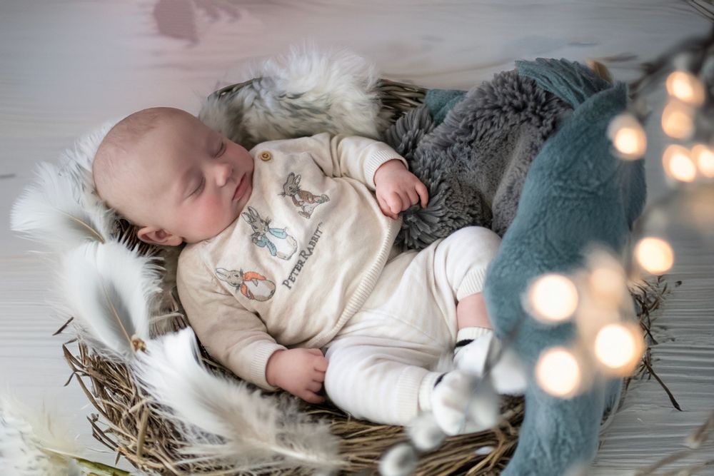 A baby sleeps peacefully in a wicker basket with soft white fur lining and bokeh lights in the background.