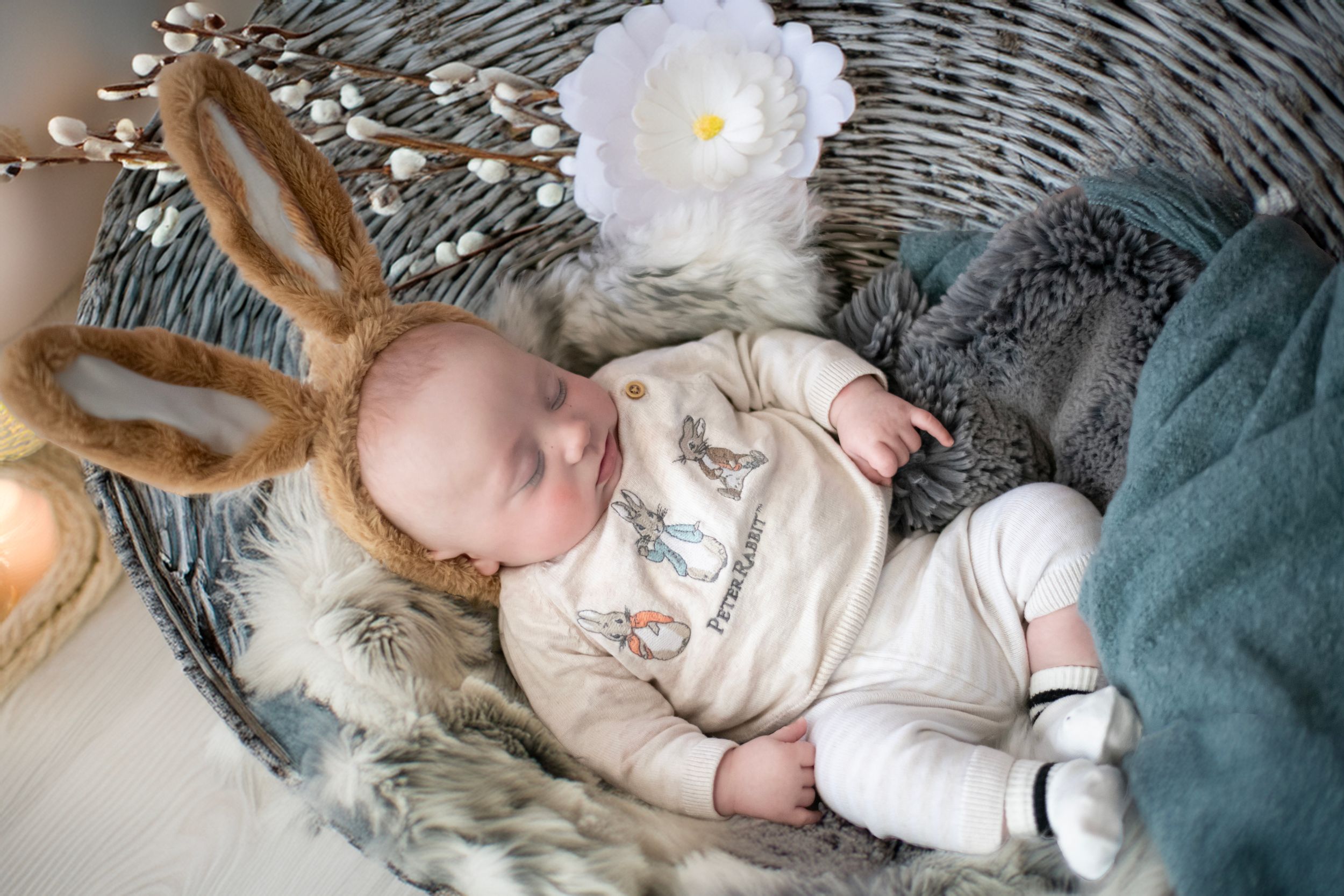 A sleeping infant rests on a gray wicker chair with decorative lights and a white floral accent.