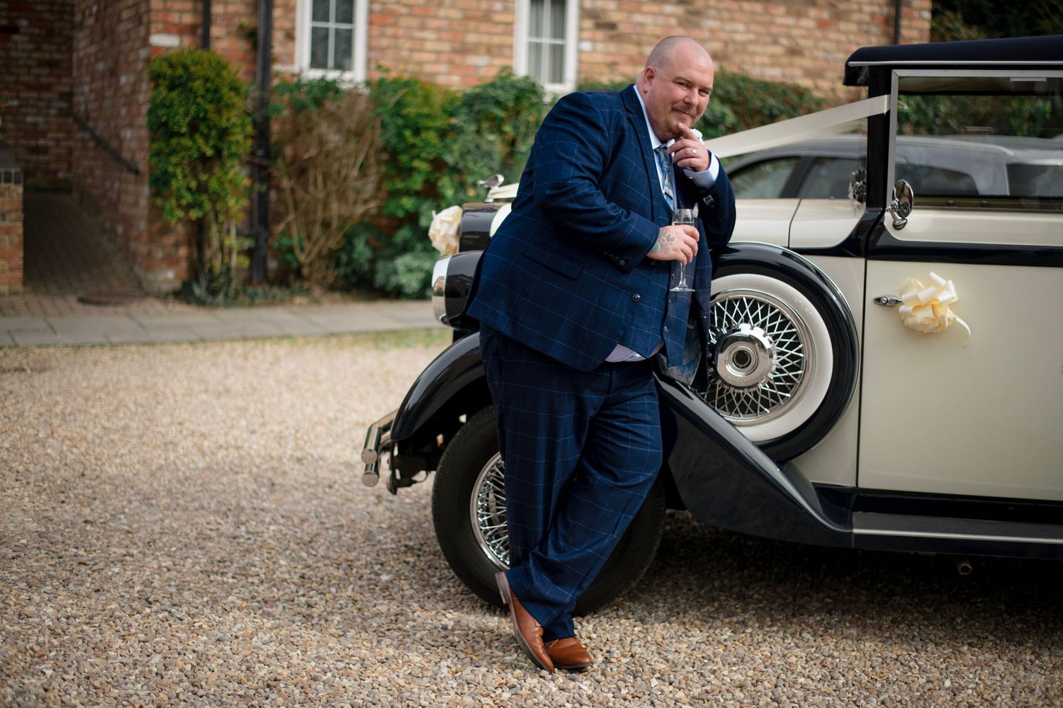 Elegant vintage car with a person in a navy suit leaning against it on a gravel driveway near a brick building.
